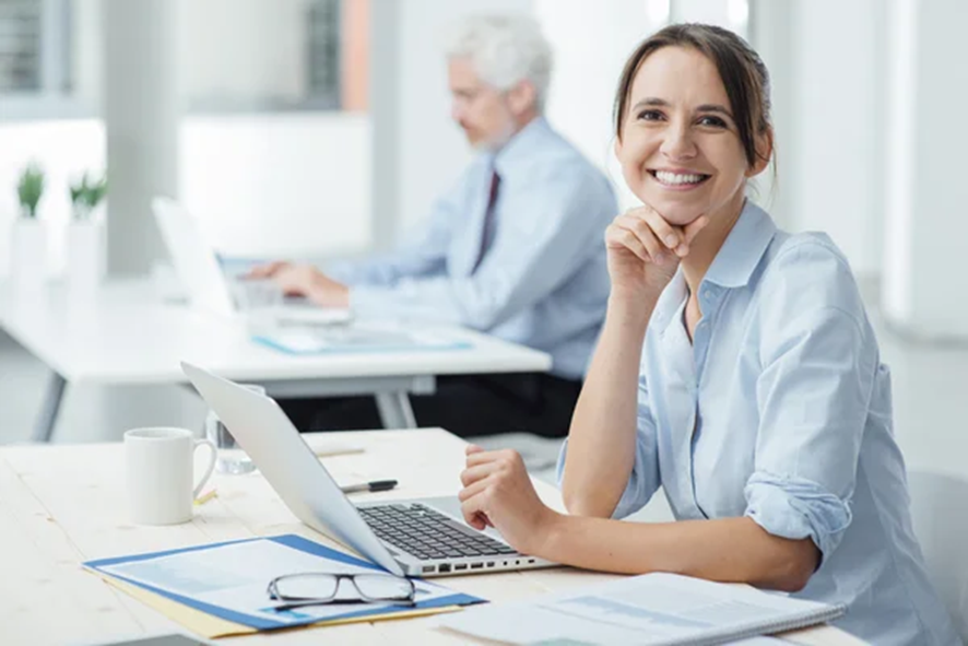 Una mujer sonriente sentada en un escritorio con una computadora portátil y apoyando la barbilla en la mano. Al fondo, un hombre trabaja en un escritorio separado.