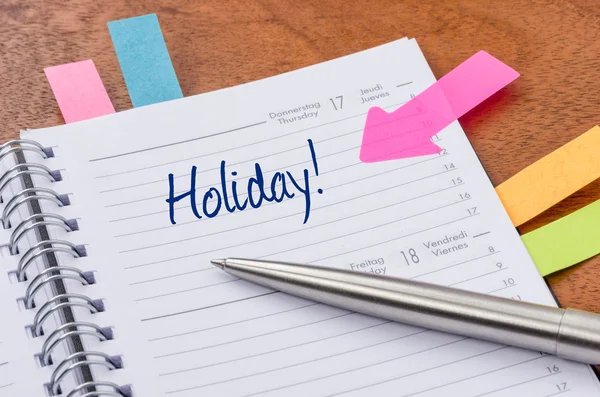 Cuaderno de espiral que muestra un calendario con pestañas de colores y la palabra "¡Vacaciones!" escrita con tinta azul, junto a un bolígrafo plateado.